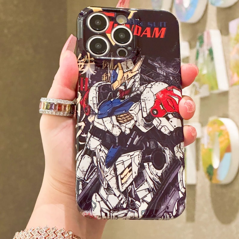 Puede incluir: Una funda de tel&eacute;fono negra con una ilustraci&oacute;n blanca, roja y azul de un robot. La funda tiene una lente de c&aacute;mara transparente con un sistema de triple lente. El texto "White Sun Gundam" est&aacute; impreso en la funda.