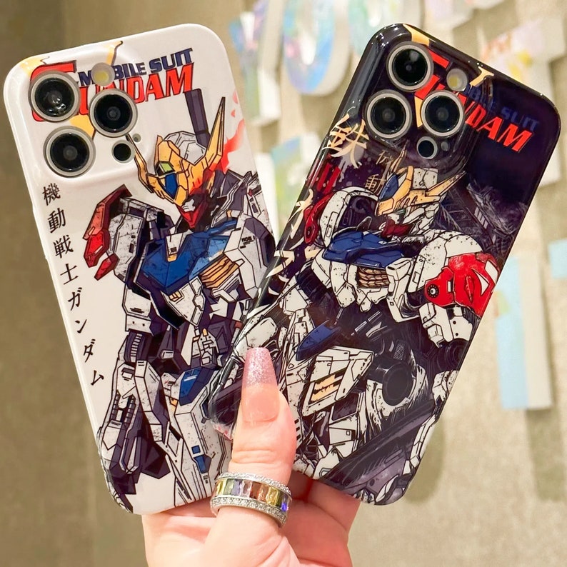 Puede incluir: Dos fundas de tel&eacute;fono blancas con una ilustraci&oacute;n en blanco y negro de un robot. El texto "Mobile Suit Gundam" est&aacute; impreso en las fundas. Las fundas tienen varios lentes de c&aacute;mara en la parte posterior.