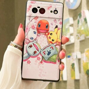 May include: A pink phone case with a black border and a design of five Pokemon characters, including Pikachu, Bulbasaur, Charmander, Squirtle, and a white cat-like Pokemon. The background is a pink and white floral pattern.