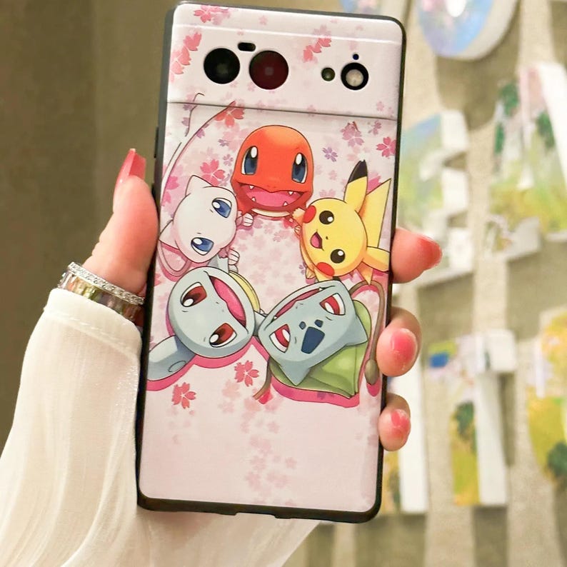 May include: A pink phone case with a black border and a design of five Pokemon characters, including Pikachu, Bulbasaur, Charmander, Squirtle, and a white cat-like Pokemon. The background is a pink and white floral pattern.