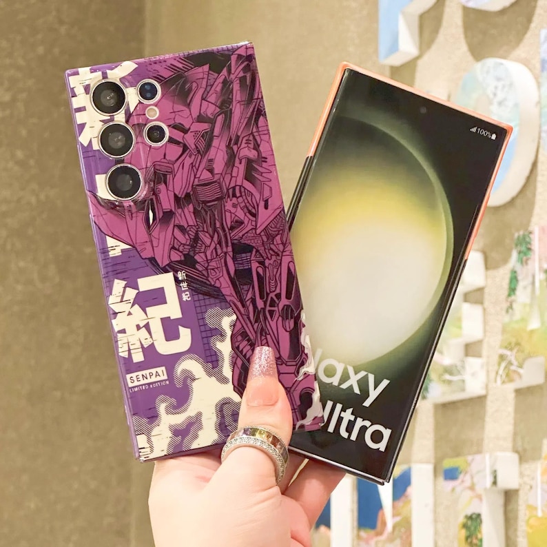 Może przedstawiać: Dwa etui na telefon, jedno z anime-stylowym wzorem w kolorze fioletowym i białym z robotem i tekstem "SENPAI LIMITED EDITION", a drugie z czarno-białym wzorem z tekstem "Galaxy Ultra".