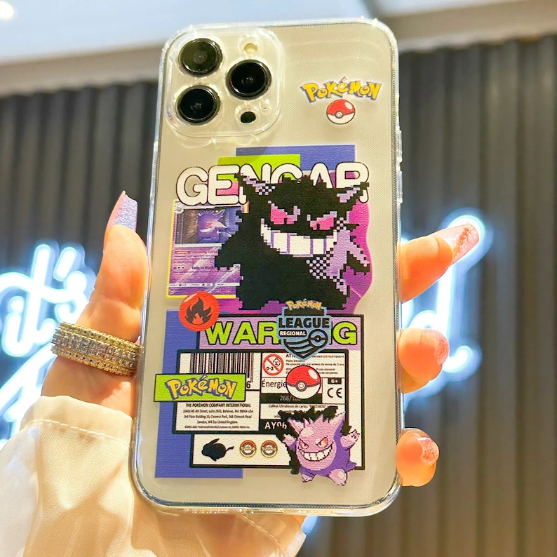 Może przedstawiać: Przezroczyste etui na telefon z motywem Pokemon. Etui ma r&oacute;żne naklejki, w tym Gengar, Pokeball i logo Ligi Pokemon. Na etui widoczny jest r&oacute;wnież tekst "WAR G".