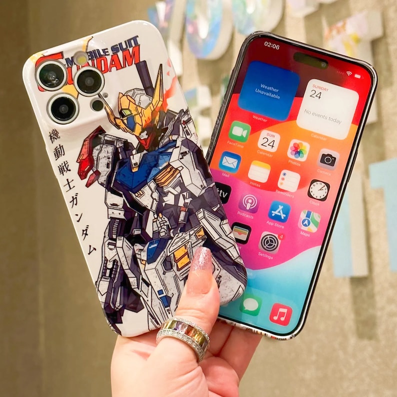 Puede incluir: Una funda de iPhone blanca con una ilustraci&oacute;n colorida de un robot Gundam de la serie de anime Mobile Suit Gundam. La funda tiene una parte posterior transparente y una protuberancia de c&aacute;mara elevada. La funda del tel&eacute;fono est&aacute; siendo sostenida por una mano que lleva un anillo.