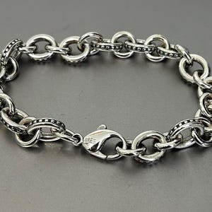 Op de afbeelding: Zilverkleurige armband met een reeks onderling verbonden, afgeronde schakels. Elke schakel is gedetailleerd met kleine, donkere accenten. De armband is beveiligd met een karabijnsluiting.