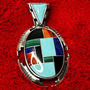 May include: A silver pendant with a geometric design. The pendant features a turquoise triangle at the top and an oval-shaped centre with a mosaic of colourful stones, including turquoise, black, orange, and green. The pendant is set against a red background.