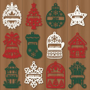 Op de afbeelding: Een set van twaalf lasergesneden kerstornamenten in rood, wit en groen. De vormen omvatten een krans met MERRY CHRISTMAS, boom met WELCOME, bal met JOY, ster met NOEL, bel met HO HO HO, kous met JOLLY, want met STAY COZY, huis met SWEET HOME, lantaarn met PEACE, sneeuwvlok met SNOW DAY, boog met HAPPY HOLIDAYS en slee met GOOD TIDINGS.