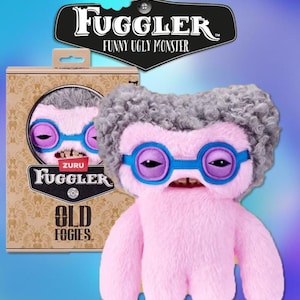 May include: A pink Fuggler plush toy with grey fur, purple and blue glasses, and a toothy grin. The toy is next to its box, which features the Fuggler logo and the words "Old Foggies."