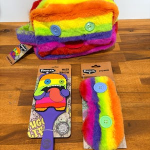Puede incluir: Un neceser, un cepillo para el pelo y una máscara para los ojos de color arcoíris. El neceser es rectangular con botones azules y verdes. El cepillo para el pelo tiene un mango morado y un diseño de monstruo colorido. La máscara para los ojos tiene botones azules y verdes.