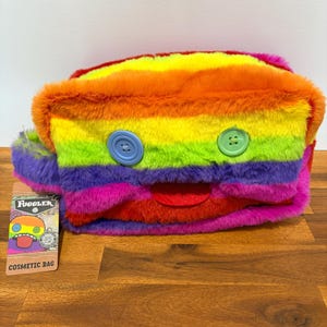 Op de afbeelding: Een regenboog gestreepte cosmetische tas met een zachte, harige textuur. De tas heeft een cartoonachtig gezicht met een rode mond en twee grote, kleurrijke knopen. Een klein label met de tekst "Fugglers" en "Cosmetic Bag" is zichtbaar.