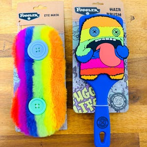 Op de afbeelding: Twee Fuggler-artikelen: een regenboogkleurig oogmasker met blauwe knopen en een blauwe haarborstel met een kleurrijk monsterontwerp. Het oogmasker heeft de tekst "EYE MASK". De haarborstel heeft de tekst "HAIR BRUSH".