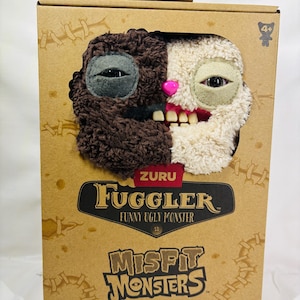 May include: A Fuggler Funny Ugly Monster toy in a cardboard box. The plush toy has a two-toned face, with one side brown and the other cream. It has mismatched eyes, a pink nose, and a toothy grin. The box reads "Misfit Monsters."