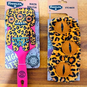 May include: A pink-handled hair brush and an eye mask. The brush features a leopard-print monster design with the word "Fuggle" at the top. The eye mask has a leopard print pattern and three eye-shaped cutouts.