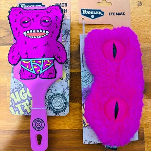 Op de afbeelding: Een roze Fuggler haarborstel en oogmaskerset. De haarborstel heeft een monsterontwerp met een brede grijns en onderbroek. Het oogmasker is pluizig met twee oogvormige uitsparingen. Beide items zitten op kartonnen verpakkingen.