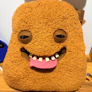 Puede incluir: Un juguete de peluche con forma de monstruo, de color canela y textura difusa, con una forma rectangular. Presenta dos ojos oscuros, brillantes y hundidos, una boca ancha con varios dientes blancos individuales y una lengua plana de fieltro rosa.