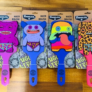 May include: Four Fuggler hair brushes with monster designs. Each brush features a different monster character with unique features, including various colours, patterns, and expressions. The brushes have a handle and are displayed on cardboard packaging.