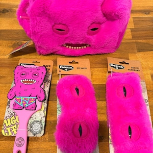 Puede incluir: Colección de accesorios Fuggler de color rosa brillante. Incluye una cara de peluche, un cepillo para el pelo, una máscara para los ojos y una diadema de belleza. Los artículos presentan el diseño del monstruo Fuggler con ojos y dientes cosidos.