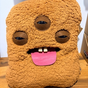 Puede incluir: Un juguete de peluche rectangular, difuso y de color tostado, con tres ojos incrustados de color marrón oscuro, dos dientes blancos prominentes y una lengua plana de fieltro rosa brillante que sobresale de una boca abierta. La textura es gruesa y rizada.