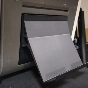 Könnte beinhalten: Ein graues und schwarzes Kunststoffgerät, das an einer grauen Maschine befestigt ist. Das Gerät hat eine flache, abgewinkelte Oberfläche und eine Lippe an der Unterseite. Das Gerät scheint ein Ständer oder Halter zu sein.