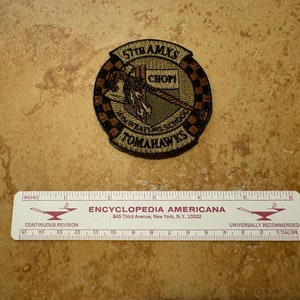 May include: A tan and brown embroidered patch with the text "SITHAMXS", "CHOPI", "1st WEAPONS SCHOOL", and "TOMAHAWKS". The patch features a stylized tomahawk with a red handle and a black and tan checkered border.