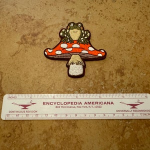 May include: An embroidered iron-on patch featuring a green frog sitting on a red and white mushroom with white dots. The mushroom has a brown stem.
