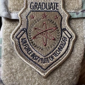 May include: Embroidered patch with the text "GRADUATE" at the top. The patch features a shield design with the words "AIR FORCE INST. OF TECHNOLOGY" around the edges. The center of the shield has a design of an atom with three stars.