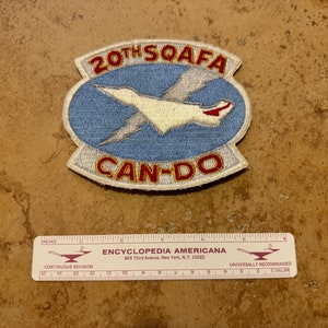 May include: A white embroidered patch with a blue background. The patch features a white airplane with a red tail, a lightning bolt, and the text "20TH SQAFA CAN-DO".