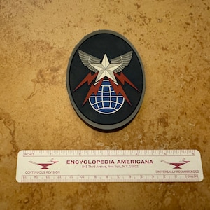 May include: A black and gray embroidered patch with a white star, red lightning bolts, and a blue globe. The patch is oval shaped and has the text "ENCYCLOPEDIA AMERICANA" and "845 Third Avenue, New York, N.Y. 10022" printed on a white ruler below it.