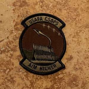 May include: A brown and green embroidered patch with a rocket and stars. The text "USAF CSOPS AIM HIGHER" is on the patch.