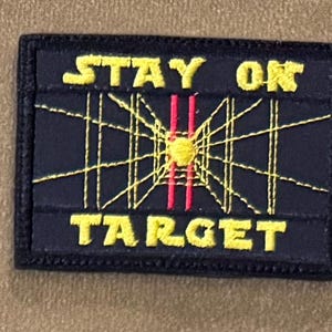 May include: Black embroidered patch with yellow stitching. The patch features a yellow starburst design with red lines in the center and the text "STAY ON TARGET".