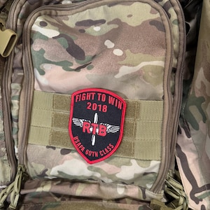 May include: A camouflage backpack with a red and black patch that says "Fight to Win 2018" and "USAF 60th Class" with a red "RTB" logo and wings.