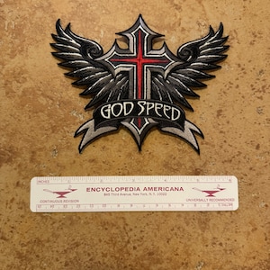 May include: A gray and red embroidered patch with a cross and wings. The patch has the words "God Speed" on a banner.