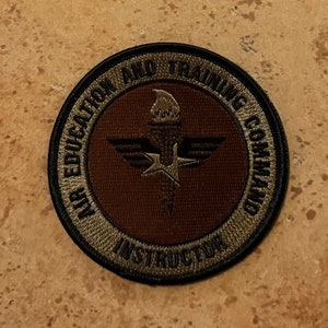May include: A green and brown embroidered patch with the text "Air Education and Training Command Instructor". The patch features a stylized torch with wings and a star.