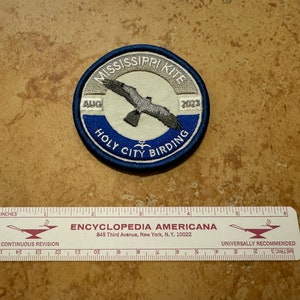May include: A round patch with a blue border features the text "MISSISSIPPI KITE" and "HOLY CITY BIRDING" around a central image of a bird. The patch includes the text "AUG 2023". A ruler with inches is below the patch.