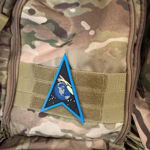 May include: A blue and black triangular patch with a white outline. The patch features a rocket launching from Earth, with a blue and white globe and a white star field. The patch is attached to a brown camouflage backpack.