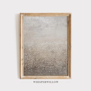 May include: A framed abstract art print in a neutral colour palette. The artwork features a gradient of grey tones with a textured appearance and a pattern of small, white dots. The frame is a light, warm gold colour, and the text "WHISPERWILLOW" is at the bottom.