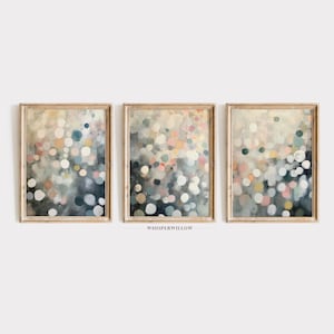 May include: Set of three framed abstract art prints. Each print features a soft, blurred design of colorful dots in shades of blue, pink, yellow, and white against a muted background. The frames are a light wood color.