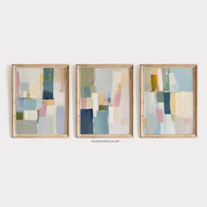 May include: Set of three abstract paintings in a minimalist style. The paintings feature geometric shapes in shades of blue, green, yellow, pink, and white. The paintings are framed in a light wood frame.
