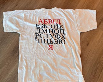 以下が含まれることがあります： グリッド形式でロシア語のアルファベットがプリントされた白い半袖Tシャツ。文字は行ごとに配置されており、最初の行と最後の文字は赤色で、その他の文字は黒色で印刷されています。