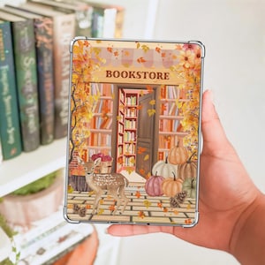 May include: A clear phone case featuring a fall-themed bookstore scene. The design includes a deer, pumpkins, and falling leaves. The word "BOOKSTORE" is displayed above a doorway filled with bookshelves. The case is held in a hand.