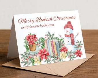 May include: A white greeting card with the text Merry Bookish Christmas to my favorite book lover. The illustration features a snowman in a red hat and scarf, a stack of colorful books, red and white poinsettias, and green pine branches.