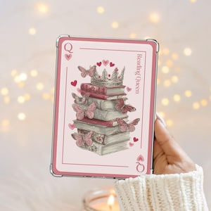 Puede incluir: Una carta de juego rosa con un diseño de libros, una corona, mariposas y corazones. La carta dice "Reading Queen" y tiene el símbolo de la Reina de Corazones. La carta está sostenida por una persona.