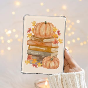 Può includere: Una custodia trasparente per tablet con un'illustrazione ad acquerello di una pila di quattro libri nei toni del marrone e dell'abbronzatura, sormontata da una grande zucca arancione e una più piccola alla base, circondata da foglie autunnali.
