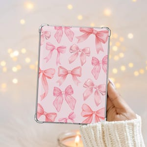 Pode incluir: Uma capa transparente com um padrão de laços rosa em aquarela sobre um fundo rosa claro. Os laços variam em tamanho e estilo, criando um design delicado e feminino. A capa é segurada em uma mão, com um fundo suave e desfocado.
