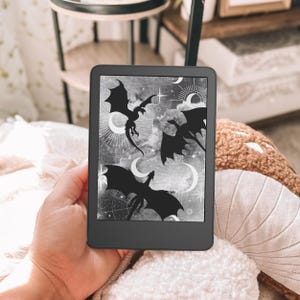 May include: A black e-reader device featuring a screen display with a grayscale graphic of three flying dragons silhouetted against a background of celestial elements, including crescent moons, stars, and radiating sunburst patterns.