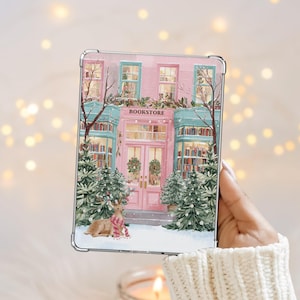 May include: A clear case featuring a festive bookstore illustration. The pink building has teal accents, Christmas trees, and a deer wearing a scarf. The word "BOOKSTORE" is above the entrance.