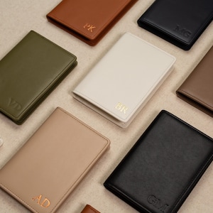 May include: An assortment of personalized leather wallets in various colors, including brown, black, olive green, and cream. Each wallet is embossed with initials in gold or silver, showcasing a minimalist design.