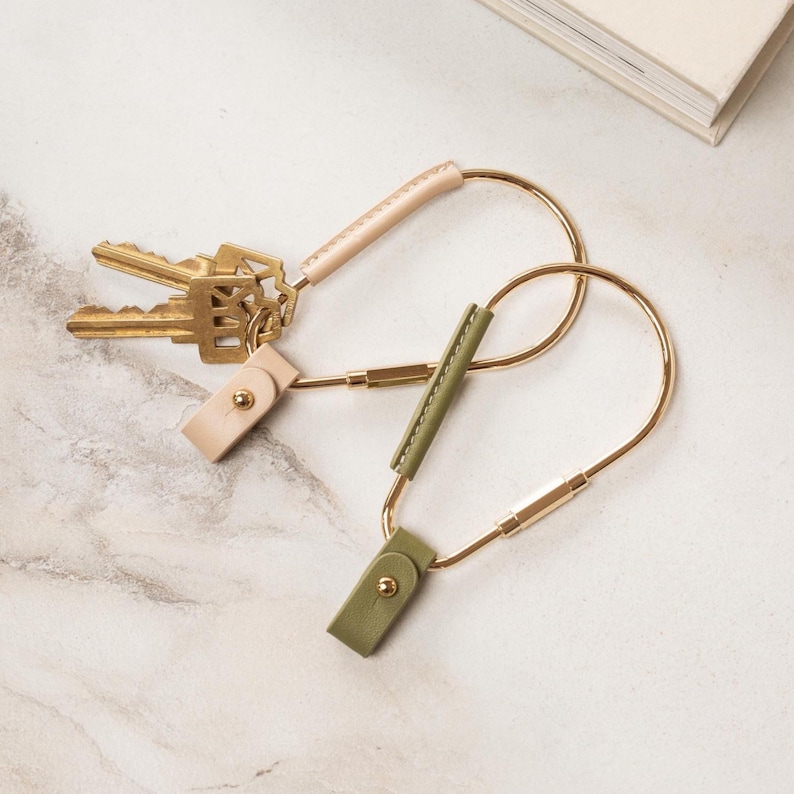 May include: Two keychains with gold-colored metal loops and leather accents. One keychain has a light pink leather accent, and the other has a green leather accent. A set of gold keys is attached to the pink keychain. The keychains are on a marble surface.