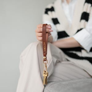 May include: A brown leather keychain with the name "VICTORIA" in gold lettering. The keychain has a gold-colored clasp and holds several keys. The leather strap is held up by a person.