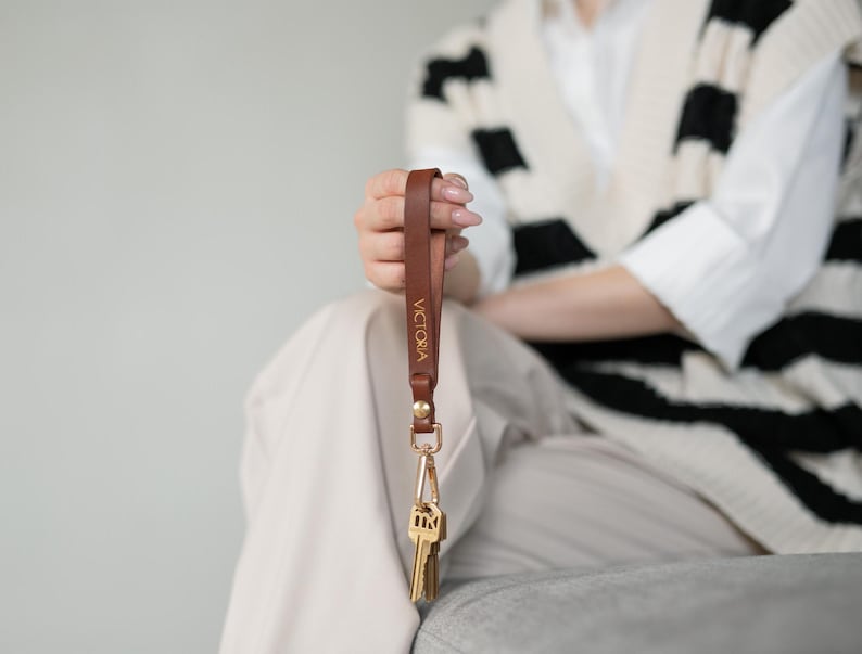 May include: A brown leather keychain with the name "VICTORIA" in gold lettering. The keychain has a gold-colored clasp and holds several keys. The leather strap is held up by a person.
