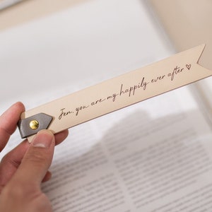 May include: A beige leather bookmark with a gray leather tab and a gold metal rivet. The bookmark has the text "Jen, you are my happily ever after" written in cursive with a small heart symbol at the end.